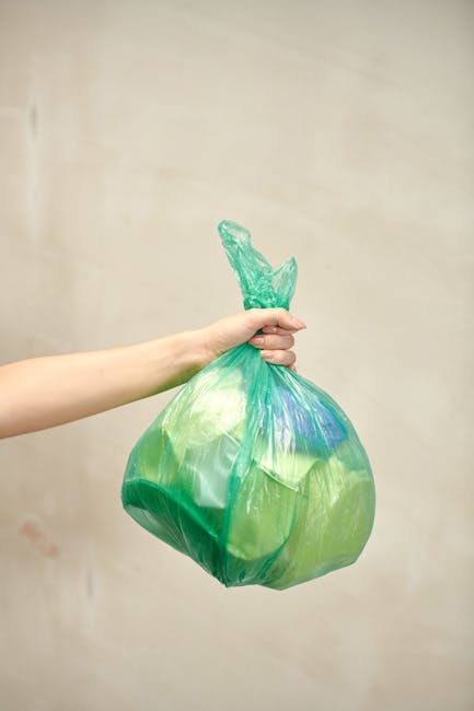 A person's arm and hand are visible, holding a semi-transparent green plastic rubbish bag by its twisted top, which is gathered and tied into a knot. The bag appears to contain mixed waste, with some visible blue and green items inside. The scene is set against a plain, light beige wall, suggesting an indoor environment, possibly a hallway or storage area. The lighting is soft and even, with no shadows or reflections. This image illustrates the act of handling waste for disposal, relevant to private rubbish collection or alternative waste handling services offered by Rubbish Collection Stockwell, emphasizing the importance of proper waste containment before collection or removal. The image’s focus on the rubbish bag and hand reinforces themes of rubbish disposal, waste management, and environmentally responsible practices.