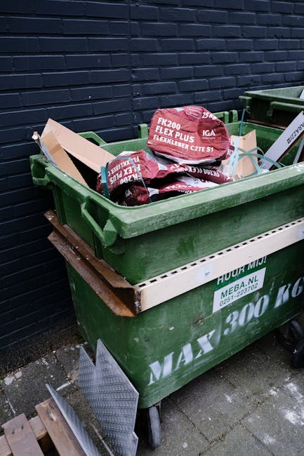 A collection of overflowing rubbish bins situated on a paved sidewalk in front of a commercial unit with a blue scaffolded facade and signage, including a yellow shop front and a sign indicating cross fish bar. The rubbish bins consist of a large grey wheeled bin filled with scattered cardboard boxes, paper, and plastic waste, with its lid partially open exposing the refuse inside. Surrounding the central bin are multiple black and red bins, some overflowing with garbage bags and assorted waste materials. Several cardboard boxes, plastic packaging, and paper are strewn on the ground around the bins, adding to the cluttered appearance. A metal railing separates the waste area from the parking space, which contains a silver hatchback car with a yellow license plate. The scene is set during daytime under diffuse natural light, with a leafless tree on the left side and part of a building with scaffolding and safety barriers visible in the background, illustrating an example of private waste accumulation that could be managed by independent rubbish removal services like Rubbish Collection Stockwell.