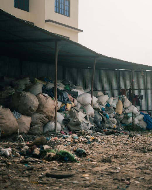 An outdoor area featuring a long row of large, irregularly shaped rubbish sacks and bags stacked against a corrugated metal fence, with some bags appearing to be made of woven plastic or thick fabric, in various shades of white, grey, and beige. The sacks are densely packed, with some overflowing or leaning, and a few smaller loads placed on top. The ground in the foreground is uneven, composed of dirt and scattered debris, including smaller pieces of rubbish and discarded materials. Above the stacks, there is a sloped metal roof supported by thin metal poles, providing limited shelter to the rubbish collection. In the background, a multi-storey building with cream-colored walls and dark-framed windows is partially visible. The atmosphere suggests a storage or temporary disposal site, possibly associated with independent waste collection services like Rubbish Collection Stockwell, highlighting an example of private or on-site rubbish storage outside residential or commercial premises, in the context of alternative waste handling and rubbish removal options.