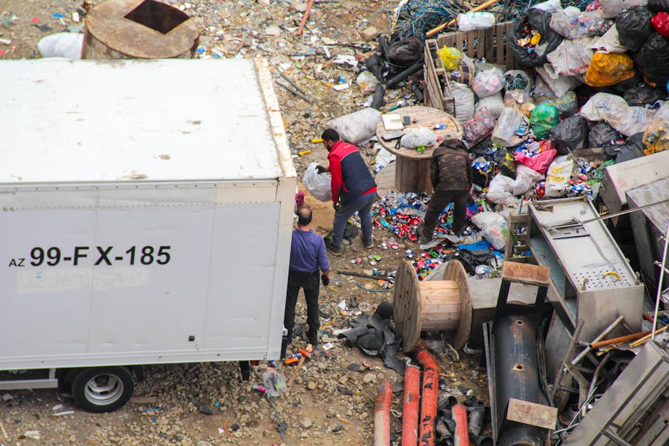 An overhead view of a cluttered outdoor area filled with various types of waste and discarded objects. Prominently, there is a large white van with black lettering displaying the code '99-FX-185' on its side, positioned on a dirt and gravel surface. Surrounding the van are three individuals actively engaged in clearing or sorting waste, with one person wearing a red and black jacket, another in dark clothing, and the third in a purple top. The ground is strewn with a mixture of trash, including plastic bottles, paper, and broken materials. To the right, there are large piles of mixed garbage bags, some shredded open, revealing their contents, alongside discarded furniture such as wooden spools, a metal frame, and pipes. The environment appears to be an open dump or waste handling site, with debris extending into the background. The scene emphasizes private waste collection or on-site clearance services, illustrating the scale of accumulated rubbish typical of non-local authority waste management efforts. Rubbish Collection Stockwell provides professional rubbish removal, as evidenced by the scene's focus on debris segregation and disposal activities.