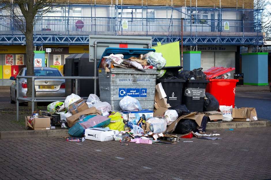 A collection of overflowing waste and rubbish bags, including black, white, and black plastic rubbish bags, as well as cardboard boxes, paper, and packaging materials, are piled on a paved area adjacent to a blue metal railing. The rubbish appears to be a mixture of household waste and discarded packaging, with some items spilling onto the street surface. Behind the pile, there is a grey metal recycling bin marked for mixed paper and cardboard, with some damaged or partially open lid causing some of the waste to be visible around it. To the left of the recycling bin, a parked silver car with a visible registration plate is positioned near a curb and a green post, while other discarded waste items are scattered around the base of the bins. In the background, a commercial building with storefronts and signage is partially covered by scaffolding and construction netting, indicating ongoing maintenance or renovation works. The scene illustrates a typical urban rubbish collection site possibly prepared for removal or awaiting collection, highlighting the need for efficient waste management services like those offered by Rubbish Collection Stockwell, especially for private or alternative waste disposal in the area.