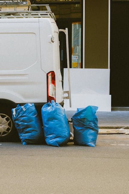 A collection of overflowing waste and rubbish bags, including black, white, and black plastic rubbish bags, as well as cardboard boxes, paper, and packaging materials, are piled on a paved area adjacent to a blue metal railing. The rubbish appears to be a mixture of household waste and discarded packaging, with some items spilling onto the street surface. Behind the pile, there is a grey metal recycling bin marked for mixed paper and cardboard, with some damaged or partially open lid causing some of the waste to be visible around it. To the left of the recycling bin, a parked silver car with a visible registration plate is positioned near a curb and a green post, while other discarded waste items are scattered around the base of the bins. In the background, a commercial building with storefronts and signage is partially covered by scaffolding and construction netting, indicating ongoing maintenance or renovation works. The scene illustrates a typical urban rubbish collection site possibly prepared for removal or awaiting collection, highlighting the need for efficient waste management services like those offered by Rubbish Collection Stockwell, especially for private or alternative waste disposal in the area.